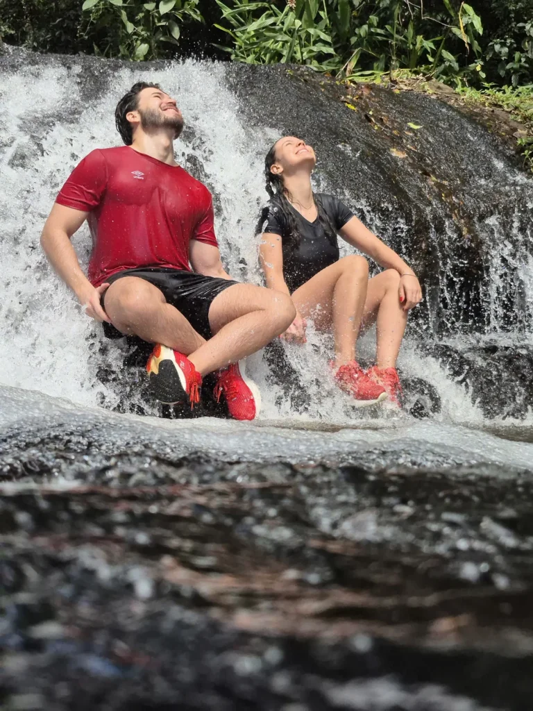 Casal sentado sob a queda d'água da cachoeira durante o passeio de quadriciclo, rindo com a água caindo nas costas.