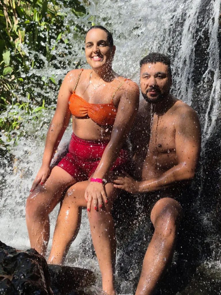 Casal sentado nas pedras, tomando banho na Cachoeira Santa Maria, sorrindo para a foto.