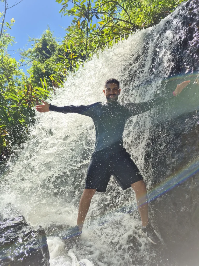 Homem de braços abertos embaixo da queda d'água da Cachoeira Santa Maria, cercado de pedras e vegetação.