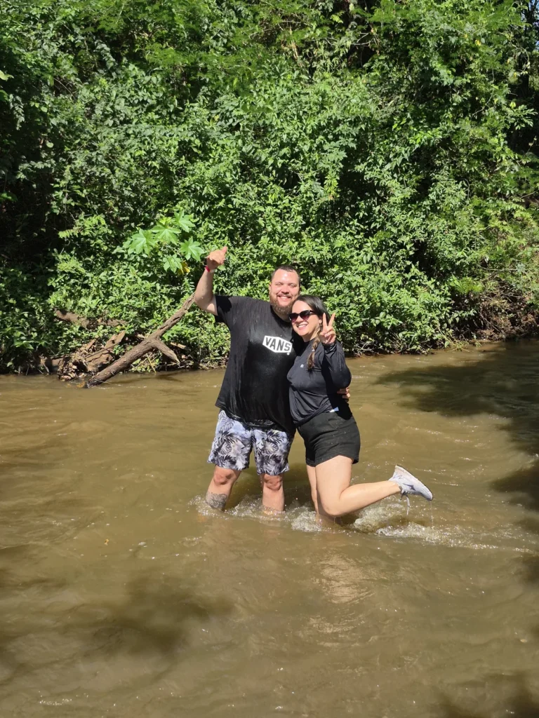 Casal brincando dentro do Rio Pinheirinho, com a água na altura dos joelhos e mata ao fundo.