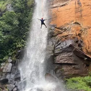 Pessoa descendo de rapel ao lado de uma cachoeira alta, em um paredão de pedra avermelhada.