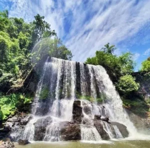 Cachoeira larga com várias quedas entre as pedras no Eco Parque Jacaré, com mata e céu azul.