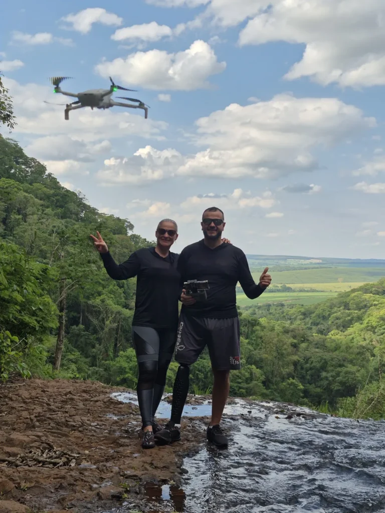 Casal no topo da Cachoeira Santa Maria, em Brotas, no passeio de quadriciclo. Ele usa prótese na perna direita, de fibra preta, e faz sinal de positivo; ela acena sorrindo. Ao fundo, mata e vale.