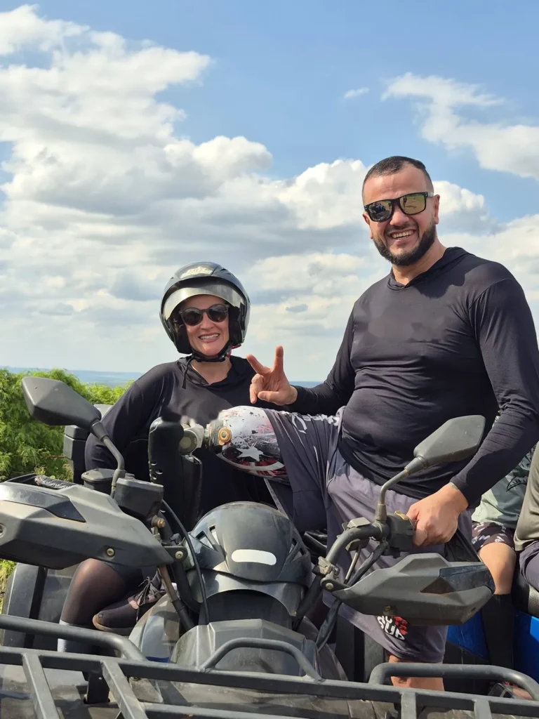 Casal no mirante da Serra Monte Cristo durante o passeio de quadriciclo. Ela está na garupa, de capacete e óculos escuros, sorrindo; ele, ao lado, apoia sobre o banco a perna com prótese estampada e faz sinal de paz.