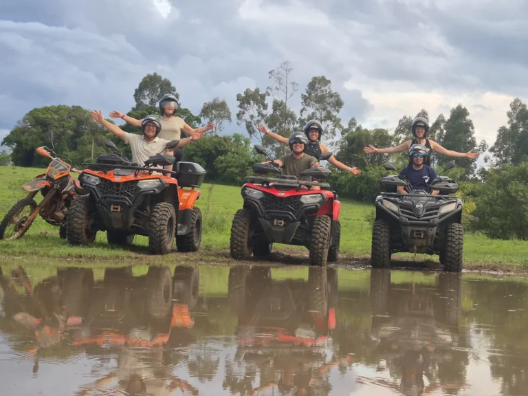 Quatro quadriciclos lado a lado em um campo, com os participantes de braços abertos e o reflexo deles numa poça d'água, no roteiro do Rio Pinheirinho.