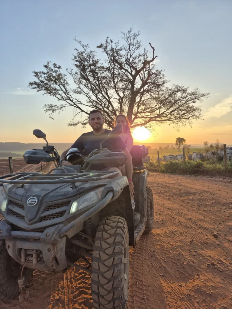 Quadriciclo parado em uma estrada de terra ao lado de uma árvore sem folhas, com o sol se pondo atrás.