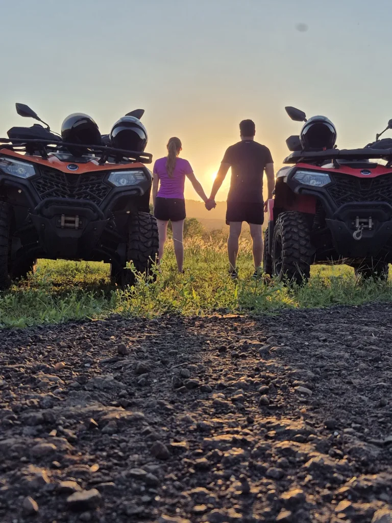 Casal de mãos dadas entre dois quadriciclos, de costas, olhando o pôr do sol em uma estrada de terra.