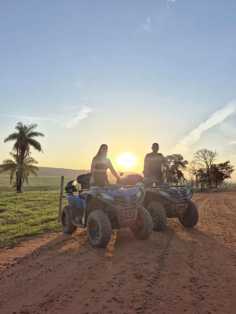 Casal sobre o quadriciclo em uma estrada de terra, com o sol se pondo atrás de uma palmeira.
