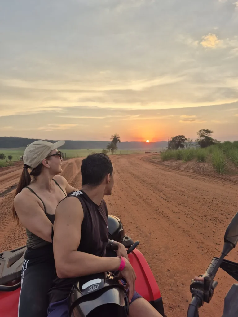 Casal no quadriciclo em uma estrada de terra vermelha, com o pôr do sol no horizonte.