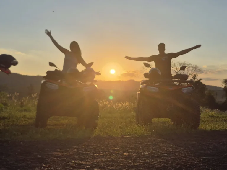 Silhuetas de duas pessoas de braços abertos sobre quadriciclos, contra o sol se pondo no horizonte.
