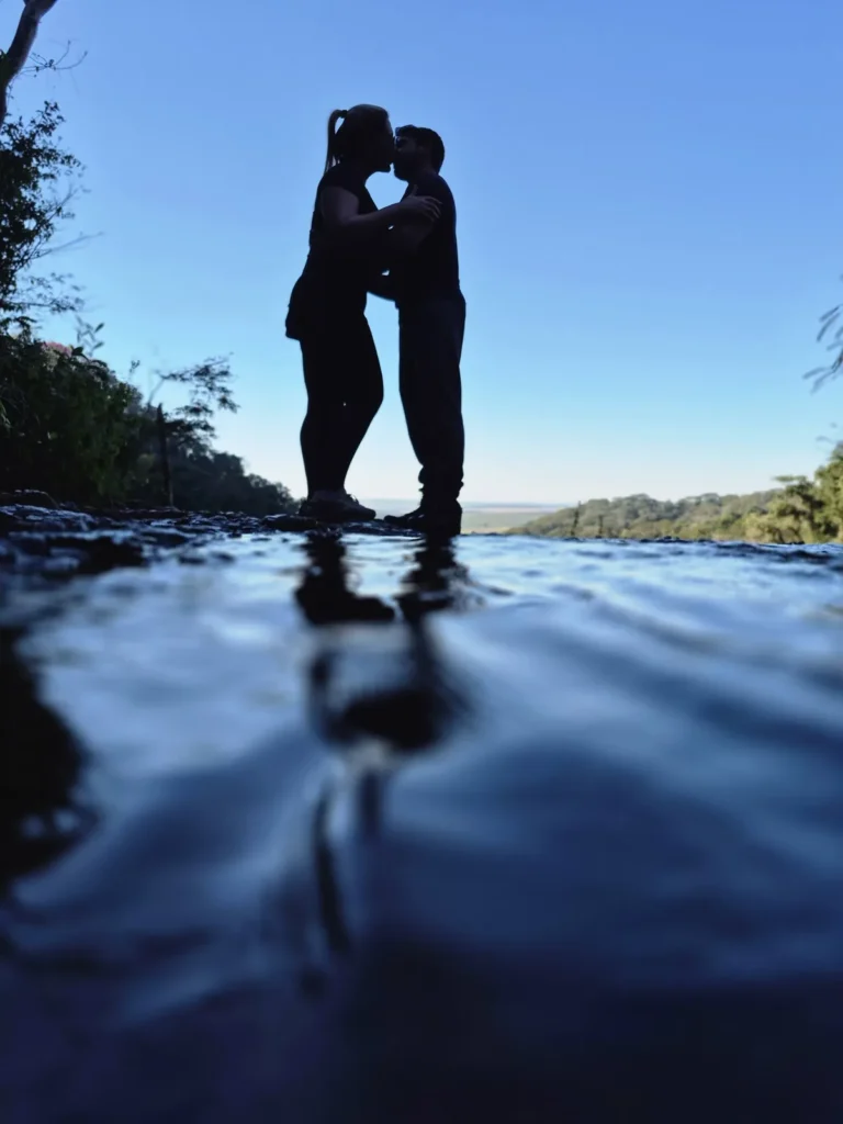 Silhueta de um casal se abraçando no topo da Cachoeira Santa Maria, refletida na água, com céu azul ao fundo.