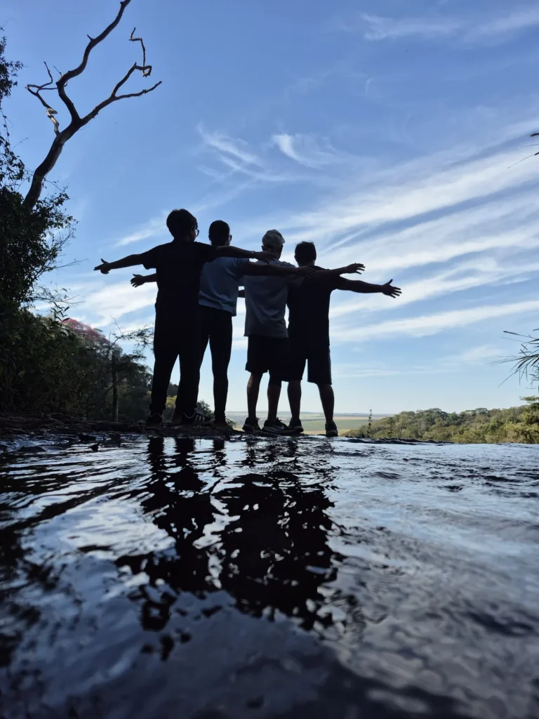 Silhuetas de um grupo de braços abertos no topo da Cachoeira Santa Maria, refletidas na água, com céu azul.