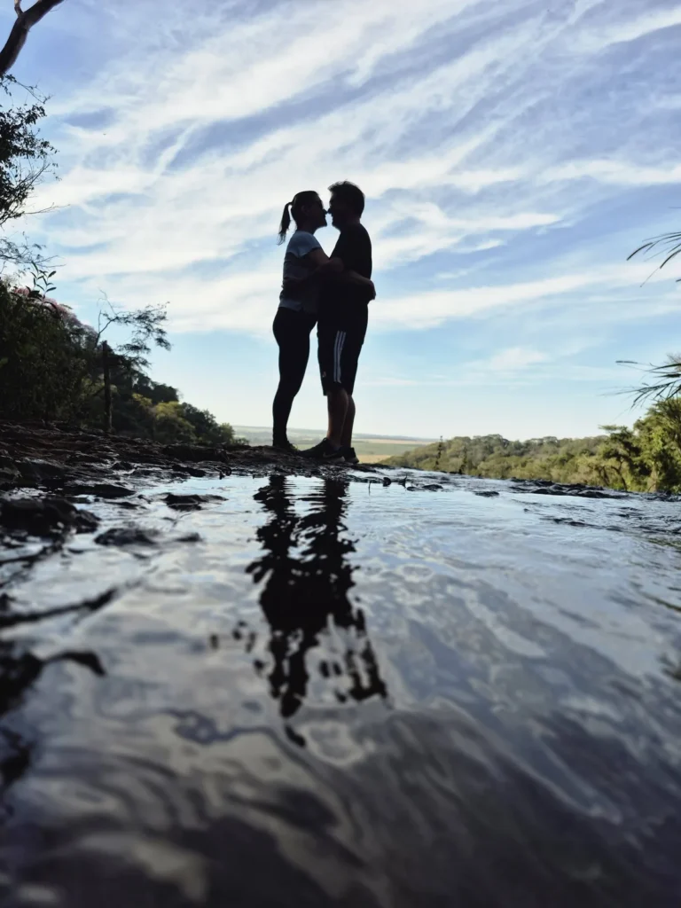 Casal no topo da Cachoeira Santa Maria, com as silhuetas refletidas na água e o céu azul com nuvens.