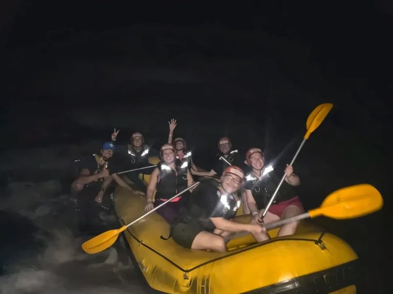 Grupo comemorando à noite em um bote de rafting amarelo, com remos para o alto.