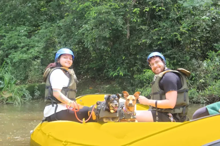 Casal de capacete e colete em um bote de rafting amarelo, com um cachorro entre eles.